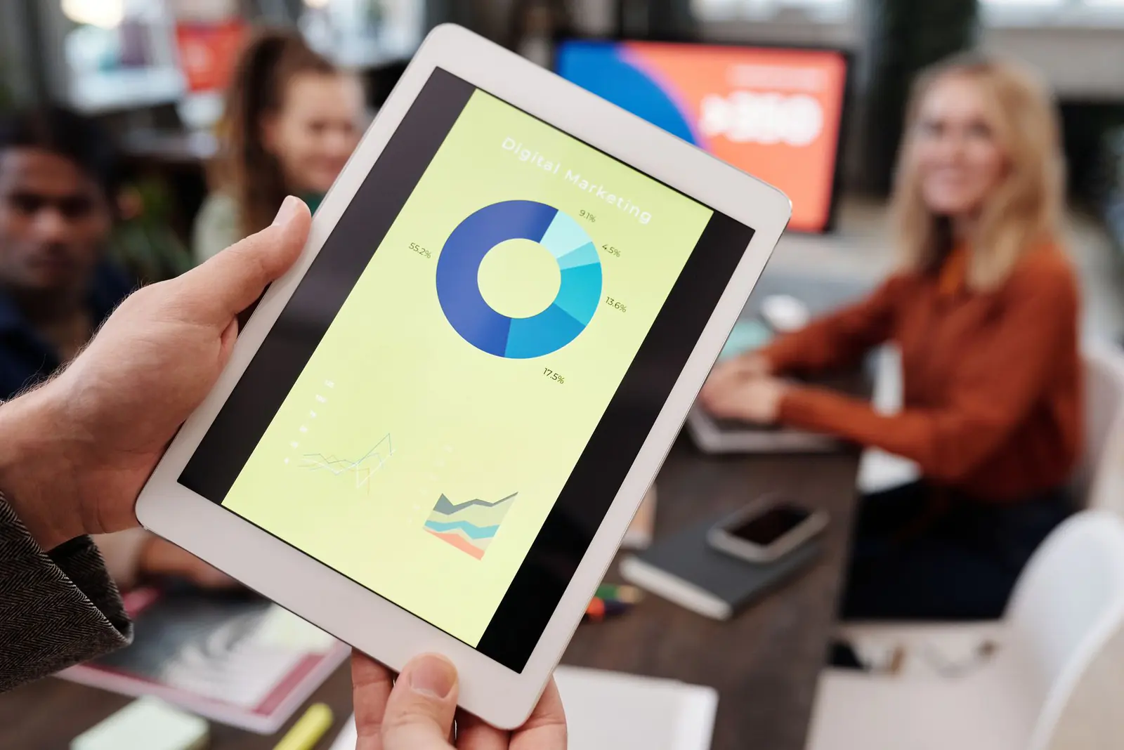 A hand holding a tablet with a report chart, a meeting behind it